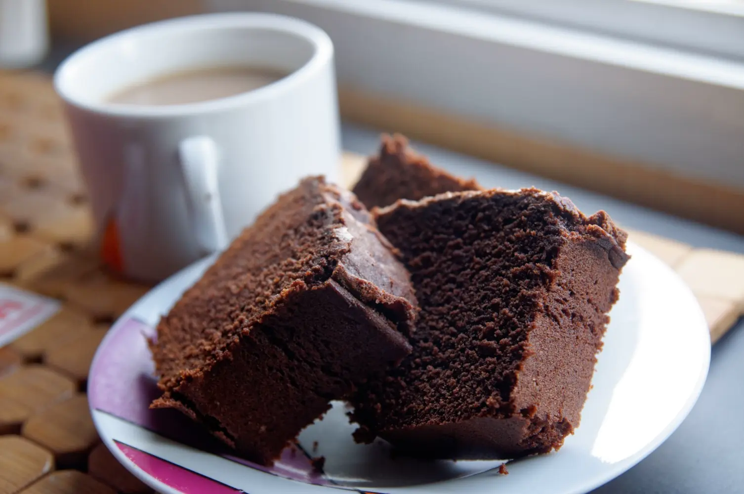 Três pedaços de bolo de chocolate com textura densa e macia servidos em um prato branco com detalhes coloridos. Ao fundo, uma xícara de café com leite complementa o cenário, criando um ambiente acolhedor, ideal para um lanche ou café da tarde.