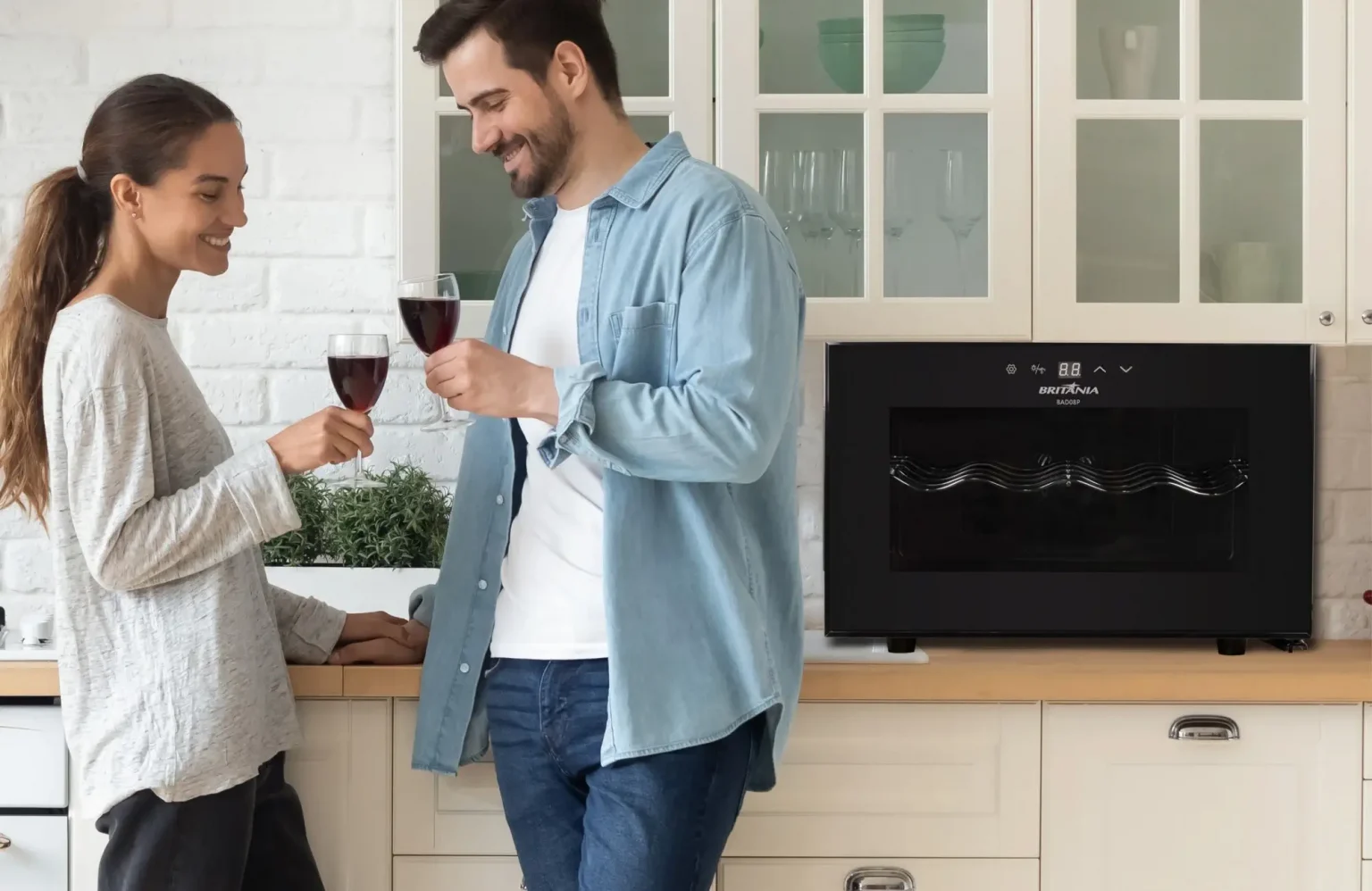 Casal brindando com taças de vinho tinto em uma cozinha moderna, com expressão de felicidade e cumplicidade. Ao fundo, uma adega climatizada Britânia sobre a bancada, com design elegante em preto, reforçando o clima aconchegante e sofisticado do ambiente.