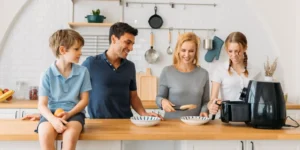 Família feliz, composta por pai, mãe, filha e filho, reunida na cozinha preparando uma refeição com uma air fryer preta. A mãe serve a comida enquanto a filha opera o aparelho, transmitindo um clima de alegria e união familiar. A cozinha tem um estilo moderno e funcional, com utensílios organizados e frutas frescas ao fundo.