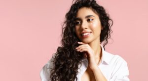 Mulher com cabelo cacheado definido, usando camisa branca e fundo rosa, tocando o rosto com expressão suave