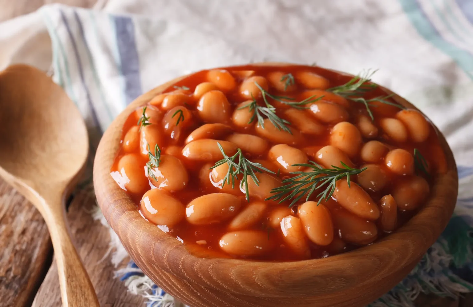 Feijão branco cozido em molho de tomate espesso, servido em uma tigela de madeira rústica e decorado com ramos frescos de dill. Ao lado, uma colher de pau e um pano de prato, compondo um cenário acolhedor e caseiro para uma refeição saborosa.
