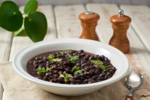 Prato fundo com feijão preto cozido e decorado com folhas de coentro ou salsa. A refeição está sobre um guardanapo de tecido claro, acompanhada de uma colher de cabo de madeira. Ao fundo, saleiro, pimenteiro e uma planta decorativa complementam o cenário rústico e acolhedor.