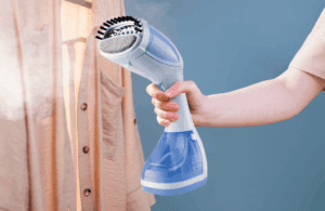 Pessoa segurando uma passadeira de roupas a vapor em um ambiente doméstico, passando vapor em uma camisa pendurada em um cabide. A passadeira é compacta, branca e azul, e está em funcionamento, emitindo vapor para desamassar e revitalizar o tecido. O fundo azul cria contraste, destacando o produto e mostrando sua praticidade para manter as roupas lisas e sem amassados.