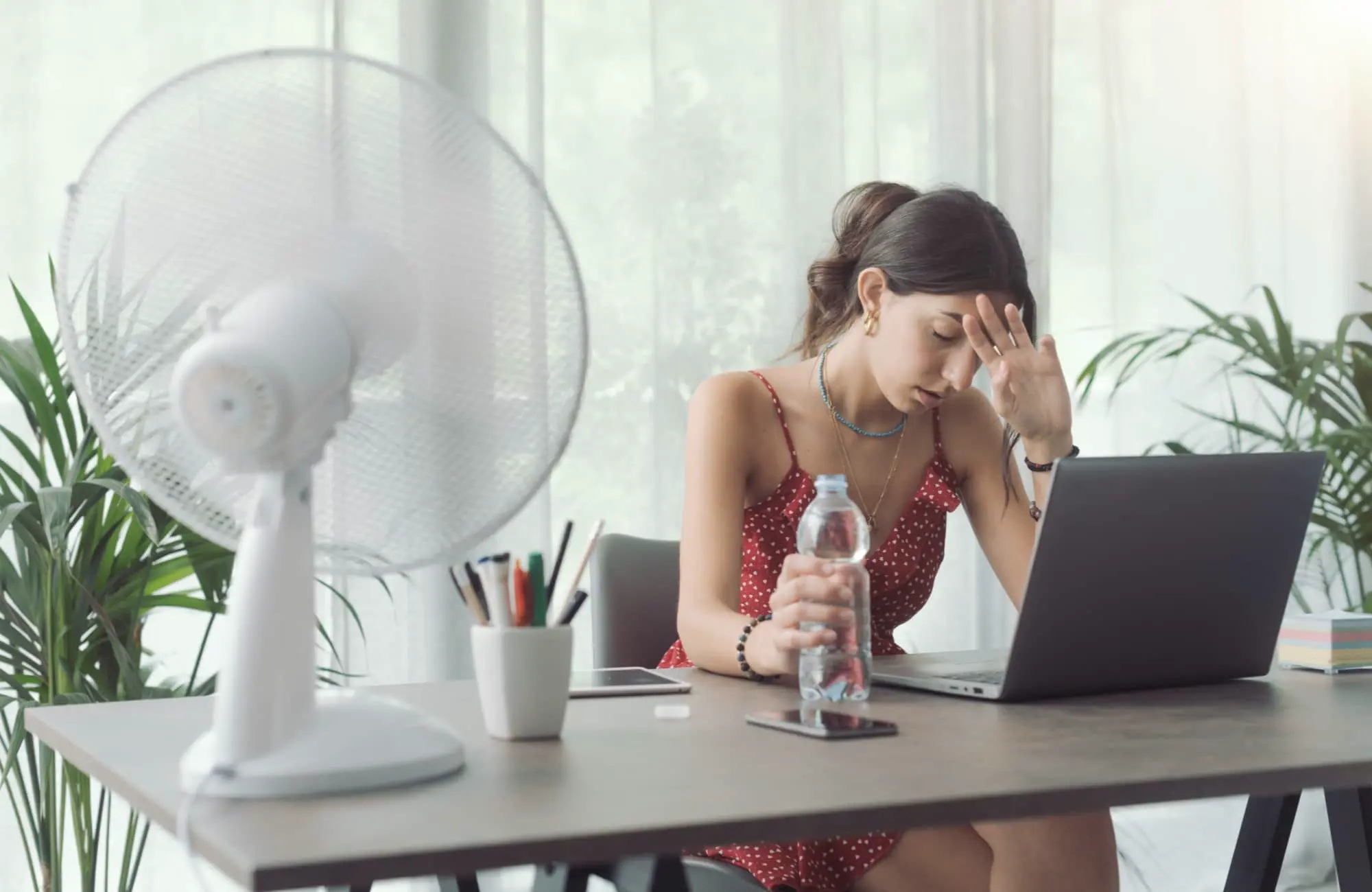Mulher jovem, sentada em uma mesa de trabalho com um ventilador ligado ao lado, segurando uma garrafa de água e demonstrando cansaço pelo calor. O ambiente é claro, com plantas ao fundo e objetos de escritório, transmitindo a sensação de um home office improvisado para lidar com as altas temperaturas do verão.