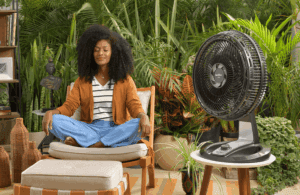 Mulher de cabelo cacheado, vestindo um cardigã laranja e calça azul, sentada confortavelmente em uma poltrona com as pernas cruzadas, olhos fechados e expressão relaxada. Ela está em um ambiente cheio de plantas, transmitindo paz e tranquilidade. Ao lado, um circulador de ar Britânia preto está ligado, proporcionando um ambiente fresco e agradável para o momento de relaxamento.