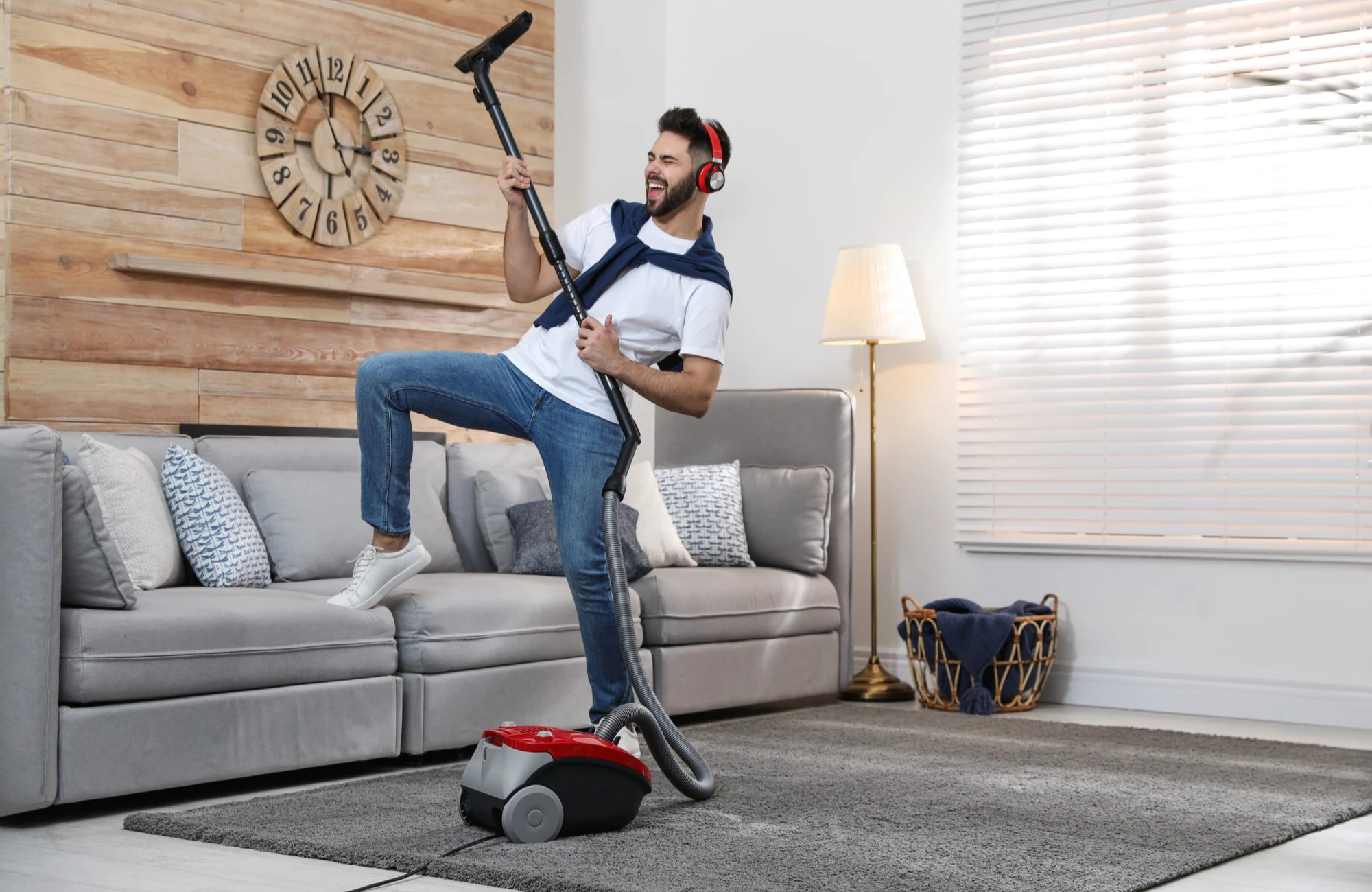 Homem jovem e sorridente, usando fones de ouvido e aspirador de pó como guitarra, em uma sala de estar aconchegante e bem iluminada. Ele se diverte enquanto limpa, mostrando que a faxina pode ser prática e até divertida com o aspirador Britânia.