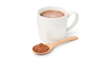 Caneca branca com chocolate quente ao lado de colher de madeira com achocolatado em pó, representando receita de chocolate quente com Nescau e leite condensado.