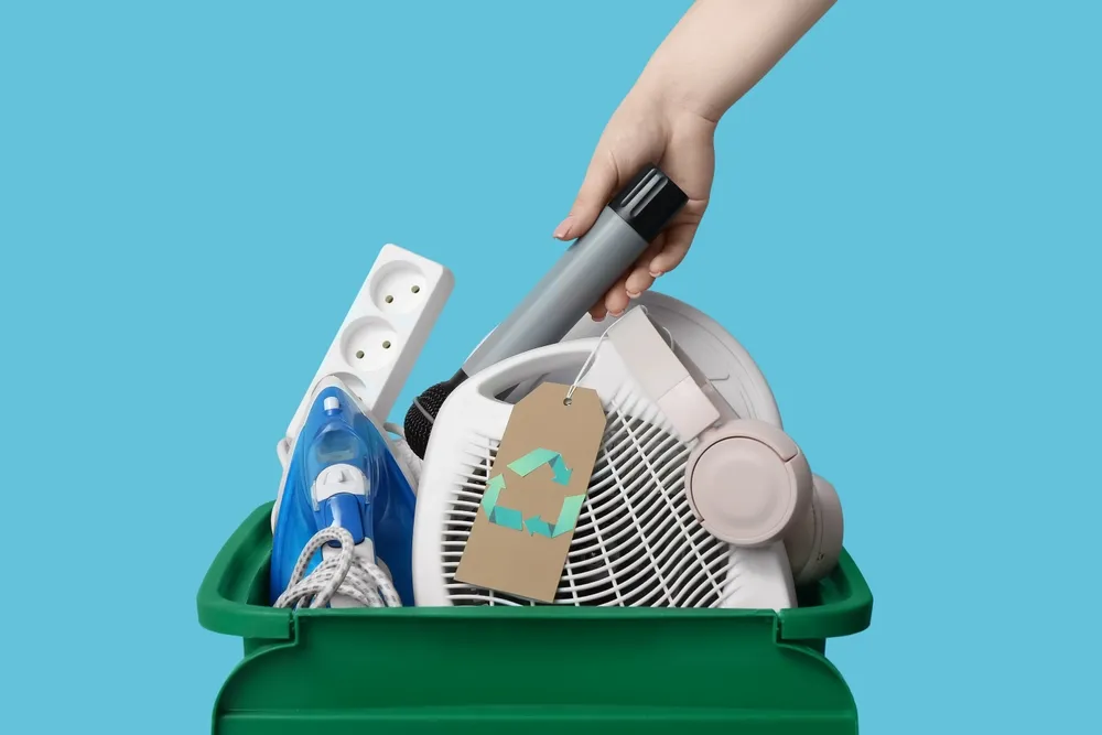Eletrodomésticos e eletrônicos usados sendo colocados em uma lixeira verde de reciclagem, representando o descarte sustentável de aparelhos domésticos.