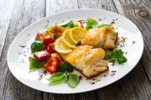 filé de bacalhau frito com legumes fritos e limão, servido em mesa de madeira.