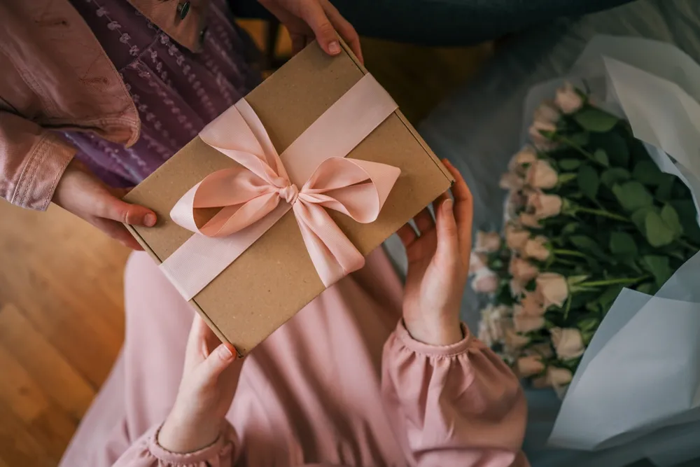 Close-up de uma criança entregando a uma mulher uma caixa de presente com uma fita rosa e um buquê de rosas delicadas sob uma suave luz natural.