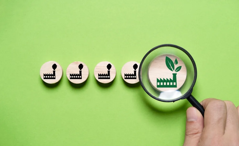 Mão segurando uma lupa e focando em uma fábrica verde em meio à produção poluente para uma economia verde, visando reduzir as emissões de dióxido de carbono conforme o Protocolo de Kyoto até 2050, em prol da sustentabilidade ambiental.