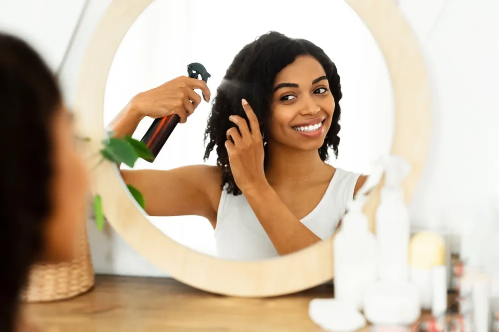 Cabeleireiro. Mulher Negra Bonita Usando Spray De Cabelo Para Fácil Pente, De Pé Perto Do Espelho Em Casa, Foco Seletivo