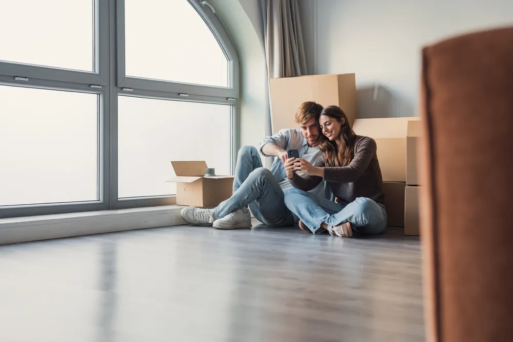 Um jovem casal ri e se abraça em sua nova casa cercada por caixas, usando um telefone celular para organizar serviços e celebrar o início de uma vida compartilhada em um novo apartamento