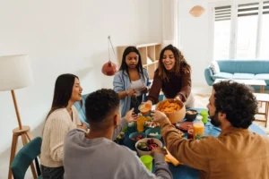 Cinco amigos diversificados, três mulheres e dois homens, sentam-se e ficam em volta de uma mesa desfrutando de lanches e bebidas juntos. Um sofá azul e mobiliário moderno melhoram o ambiente para a sua refeição casual.