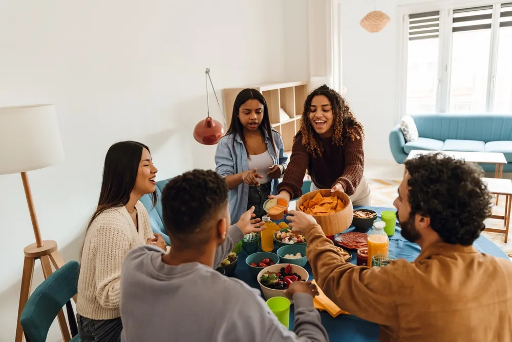Cinco amigos diversificados, três mulheres e dois homens, sentam-se e ficam em volta de uma mesa desfrutando de lanches e bebidas juntos. Um sofá azul e mobiliário moderno melhoram o ambiente para a sua refeição casual.