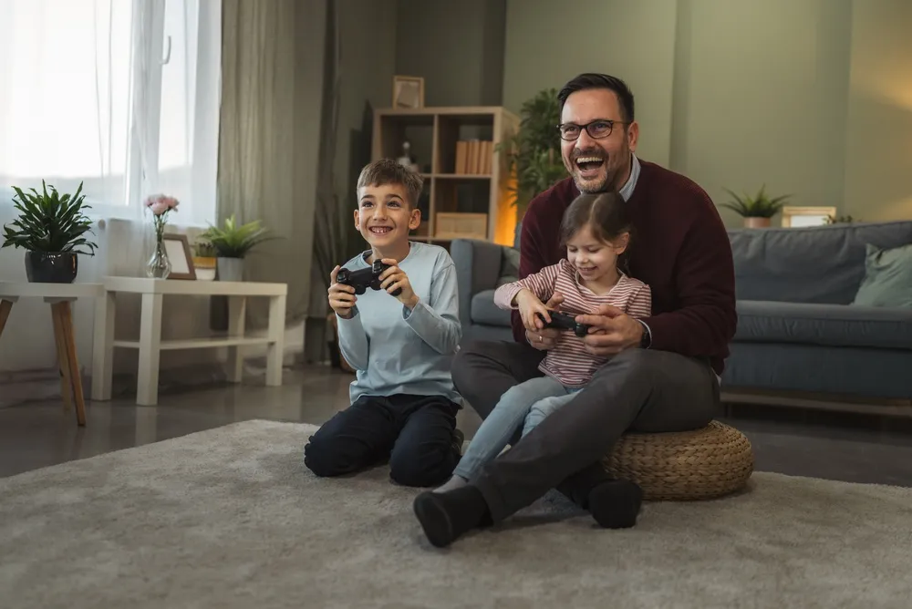 Pai e seu filho e filha estão alegremente jogando videogames juntos, compartilhando risos e laços durante o tempo em família em sua aconchegante sala de estar em casa