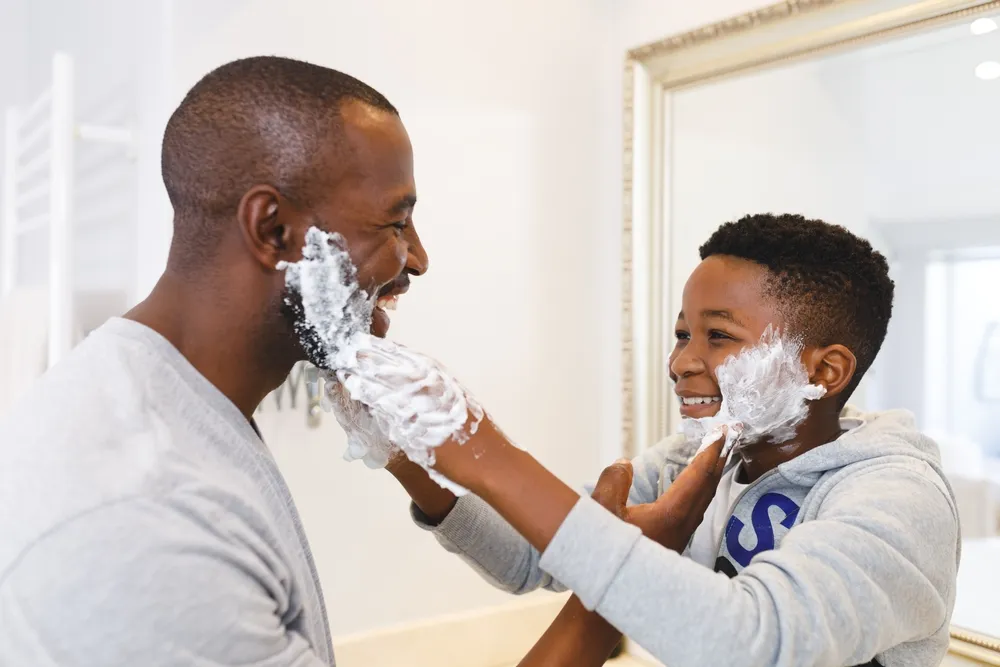 Pai afro-americano e filho se divertindo com espuma de barbear no banheiro. Família passando um tempo em casa.