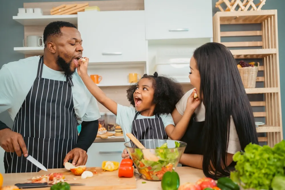 Pais afro-americanos gentis ensinando sua filha adorável a preparar comida saudável; espaço livre na cozinha; família negra feliz preparando comida saudável na cozinha juntos.