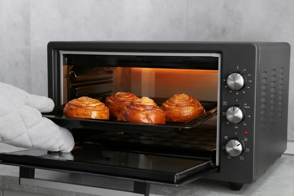 Mulher retirando do forno elétrico uma assadeira com pãezinhos em espiral deliciosos, em close