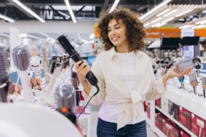 Mulher de cabelo encaracolado sorridente comparando escova de secador de cabelo e curler de cabelo, comprando produtos de estilo de cabelo na loja de eletrônicos de consumo