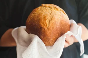Pão branco caseiro segurado pelas mãos sobre uma toalha branca com textura de waffle; avental preto.